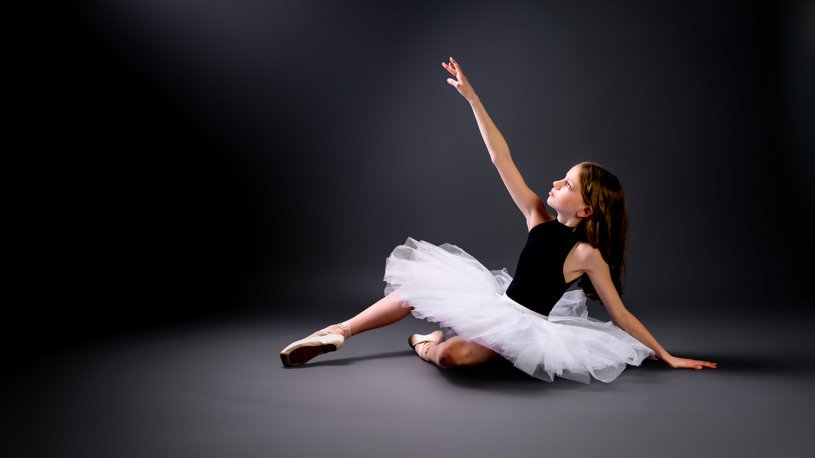 Young ballet dancer in a black leotard and white tutu reaching upward in a studio portrait
