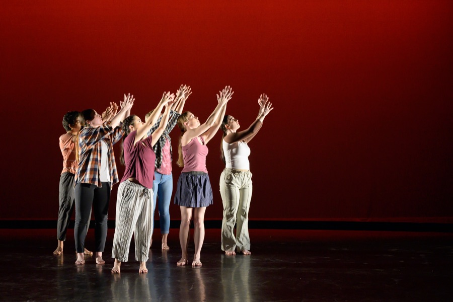 Performance group reaching upward on stage against a red backdrop