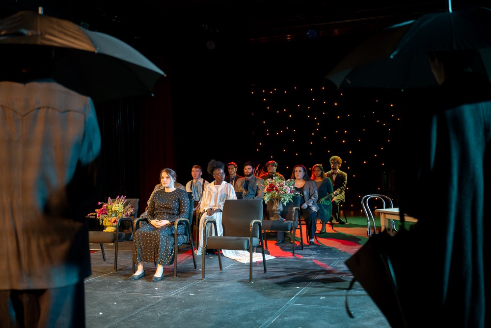 Cast seated on stage during a theatrical scene in a dark performance space